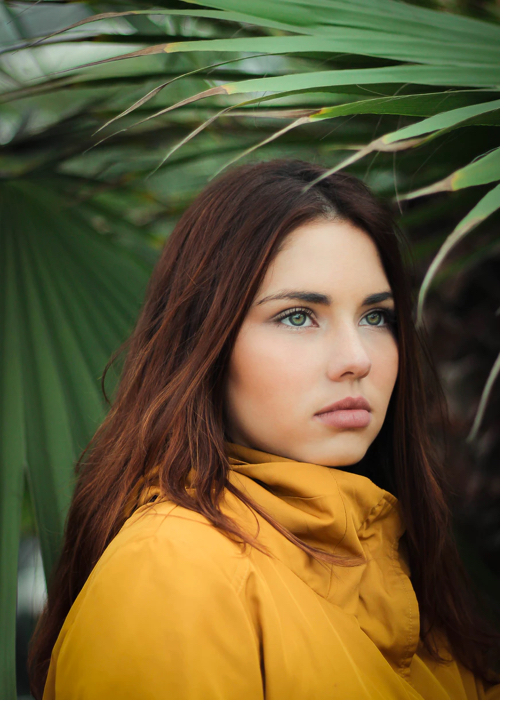 A green eyed women with brown hair standing in front of greenery wearing a yellow top.